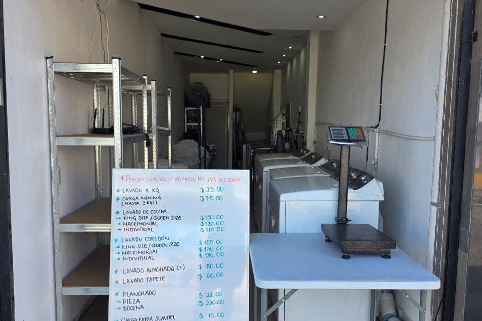 La tienda de Osoji abierta a la calle: la estantería, el pizarrón con la lista de precios, las lavadoras al fondo y la mesa con la báscula